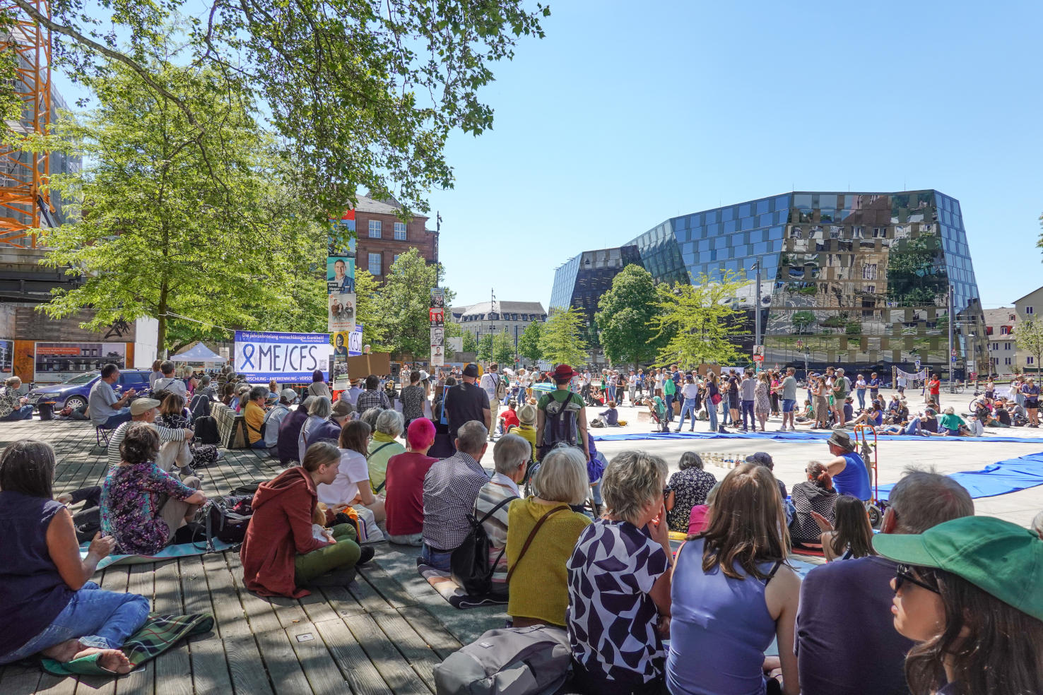 Bildbeschreibung: Foto von dutzenden Demonstrant\*innen, die aufgrund des starken Sonnenscheins auf der überschatteten Holzterrasse am Rand des Platzes sitzen.